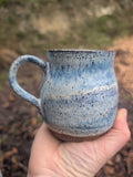 A handmade ceramic mug with a blue and white speckled glaze, held in a person's hand against a blurred outdoor background.