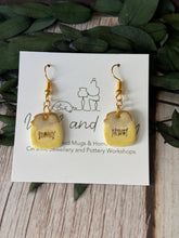 A pair of handmade dangle earrings shaped like mini toast slices, displayed on a white branded card. The earrings have a pale yellow glazed ceramic body with gold-colored metal wire hooks and a small brown Hebrew word printed on each toast.