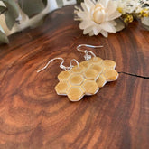 A pair of honeycomb-shaped dangle earrings with a natural tan resin body and silver fishhook wires, displayed on a rustic wooden surface with white flowers in the background.