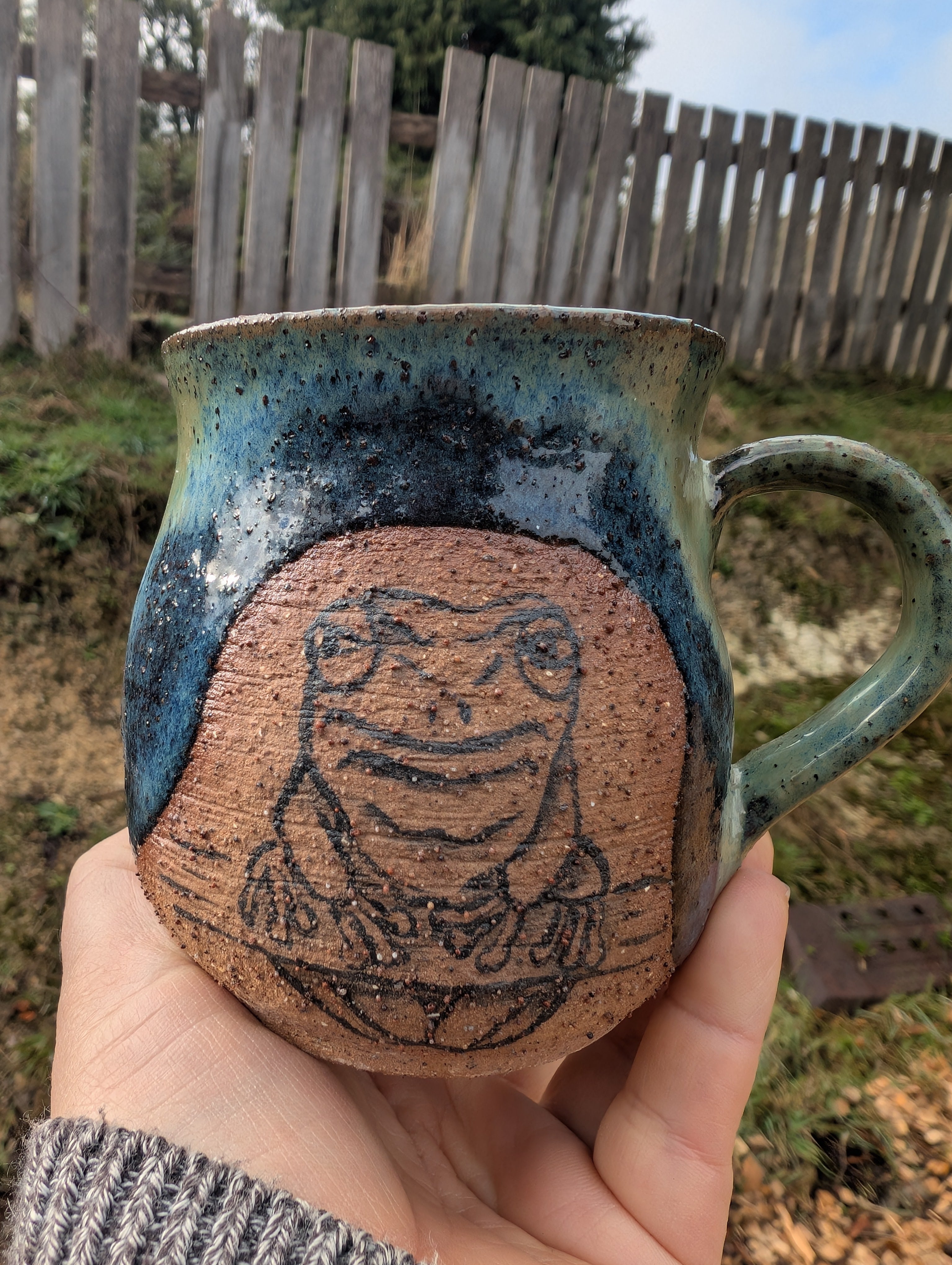 A handmade ceramic mug with a hand-drawn frog design, held in a person's hand outdoors. The mug features a speckled blue and green glaze with a textured terracotta-colored panel showing the frog sketch.