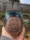 A handmade ceramic mug with a hand-drawn frog design, held in a person's hand outdoors. The mug features a speckled blue and green glaze with a textured terracotta-colored panel showing the frog sketch.