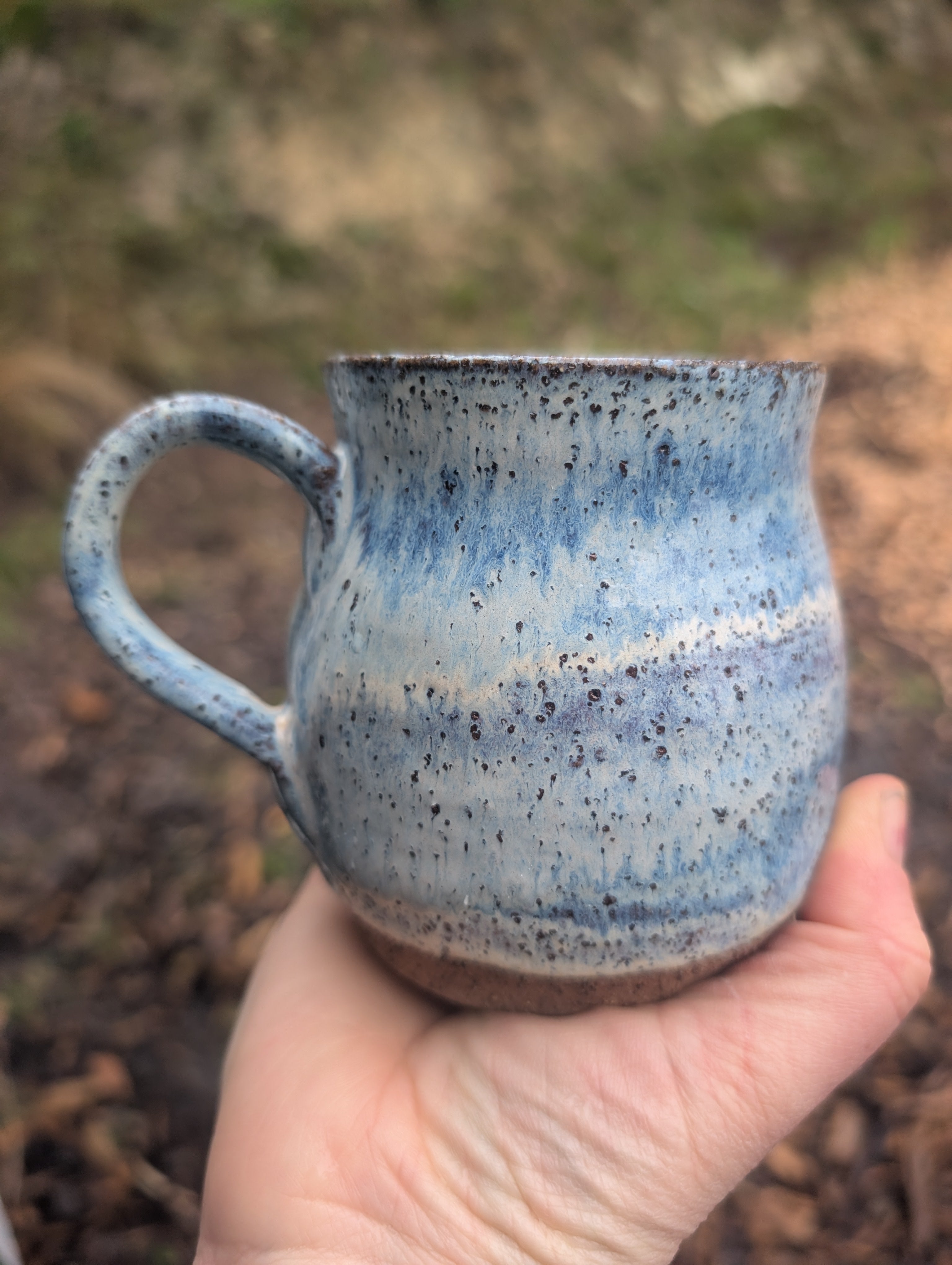 A handmade ceramic mug with a blue and white speckled glaze, held in a person's hand against a blurred outdoor background.
