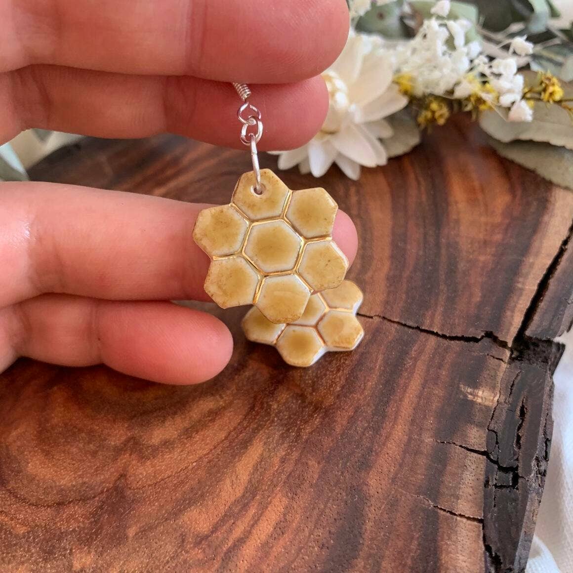 A hand holding a honeycomb-shaped pendant necklace with a silver-tone chain, set against a rustic wooden surface with dried white flowers in the background.