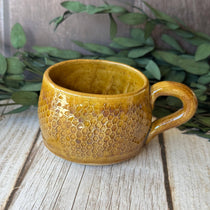 A handmade ceramic mug with a textured honeycomb pattern and a warm mustard-yellow glaze, resting on a rustic wooden surface with green foliage in the background.
