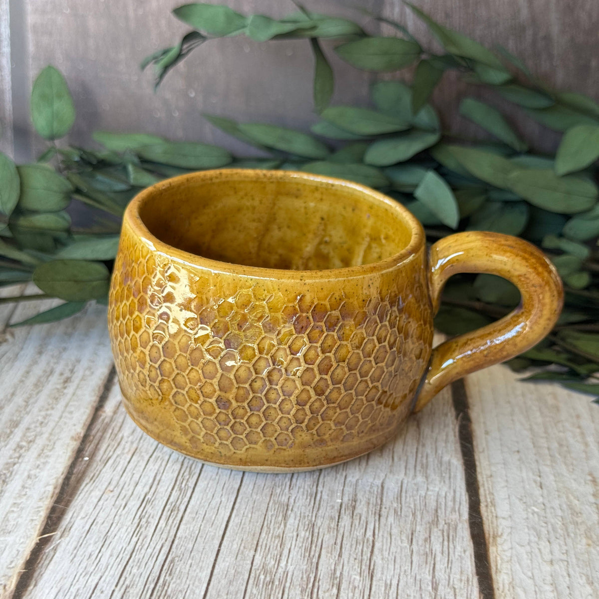 A handmade ceramic mug with a textured honeycomb pattern and a warm mustard-yellow glaze, resting on a rustic wooden surface with green foliage in the background.