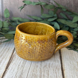 A handmade ceramic mug with a textured honeycomb pattern and a warm mustard-yellow glaze, resting on a rustic wooden surface with green foliage in the background.