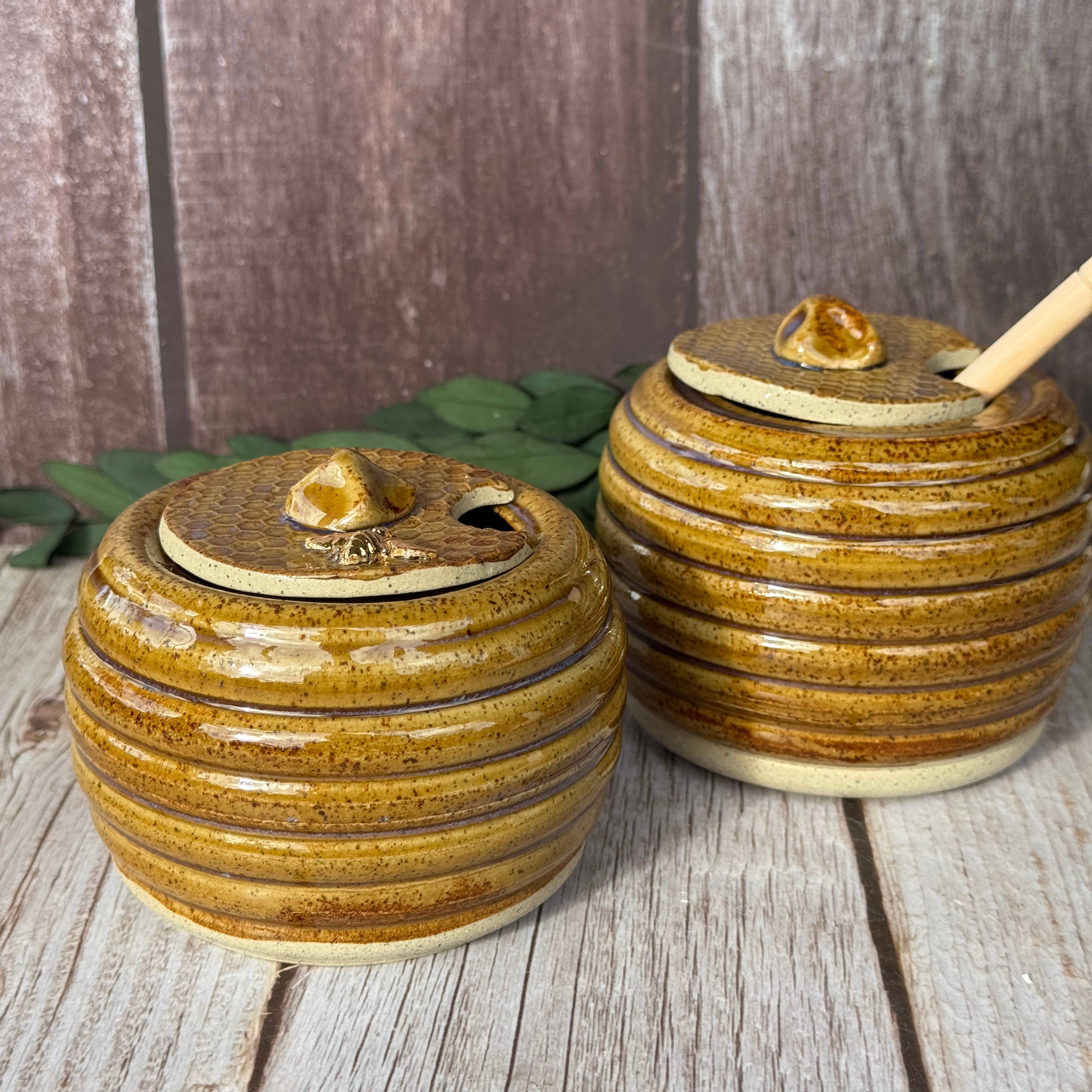 Two round ceramic honey pots with lids and wooden spoons, featuring a rustic brown and gold glaze, set on a weathered wood surface.