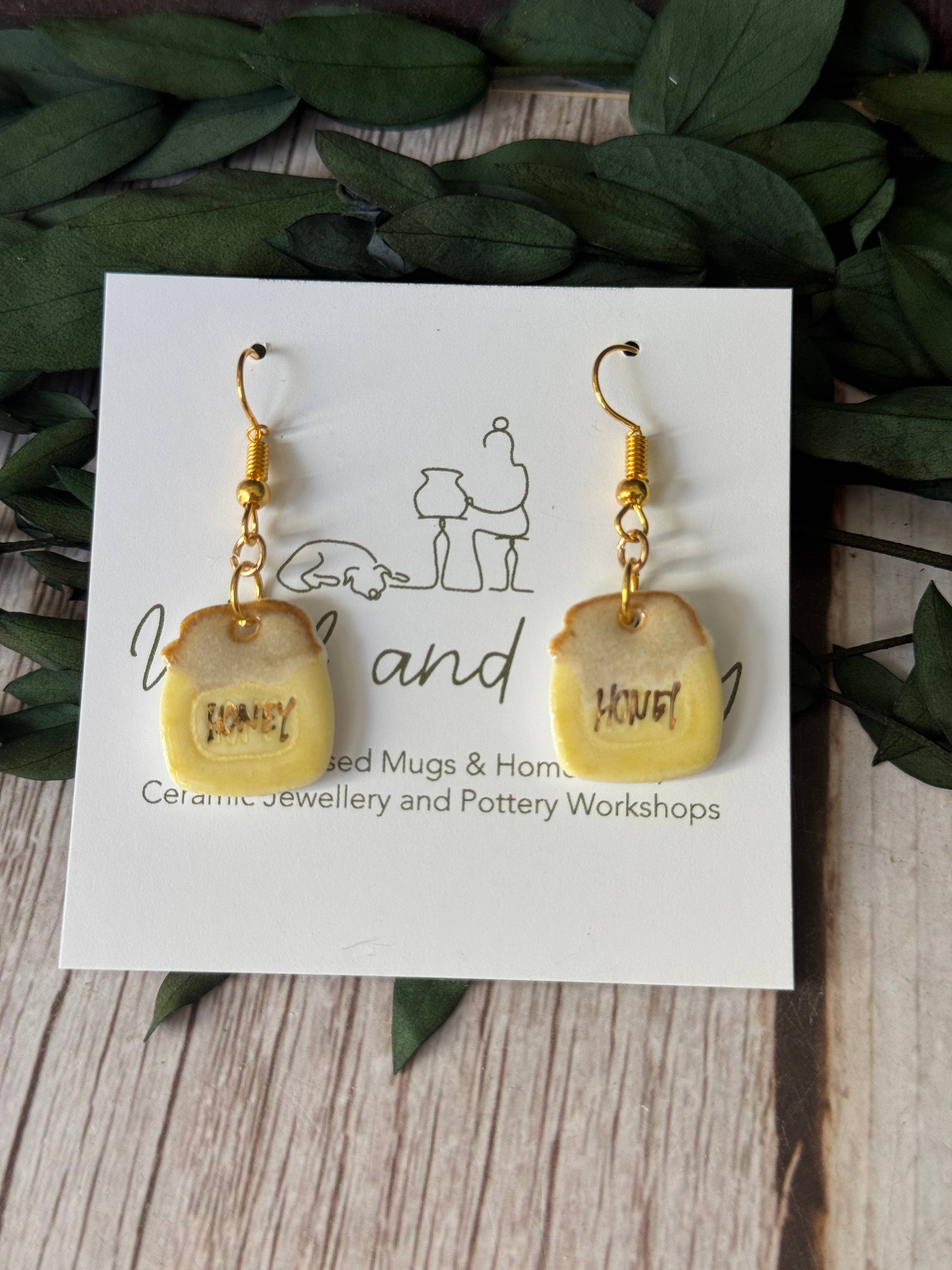 A pair of handmade dangle earrings shaped like mini toast slices, displayed on a white branded card. The earrings have a pale yellow glazed ceramic body with gold-colored metal wire hooks and a small brown Hebrew word printed on each toast.