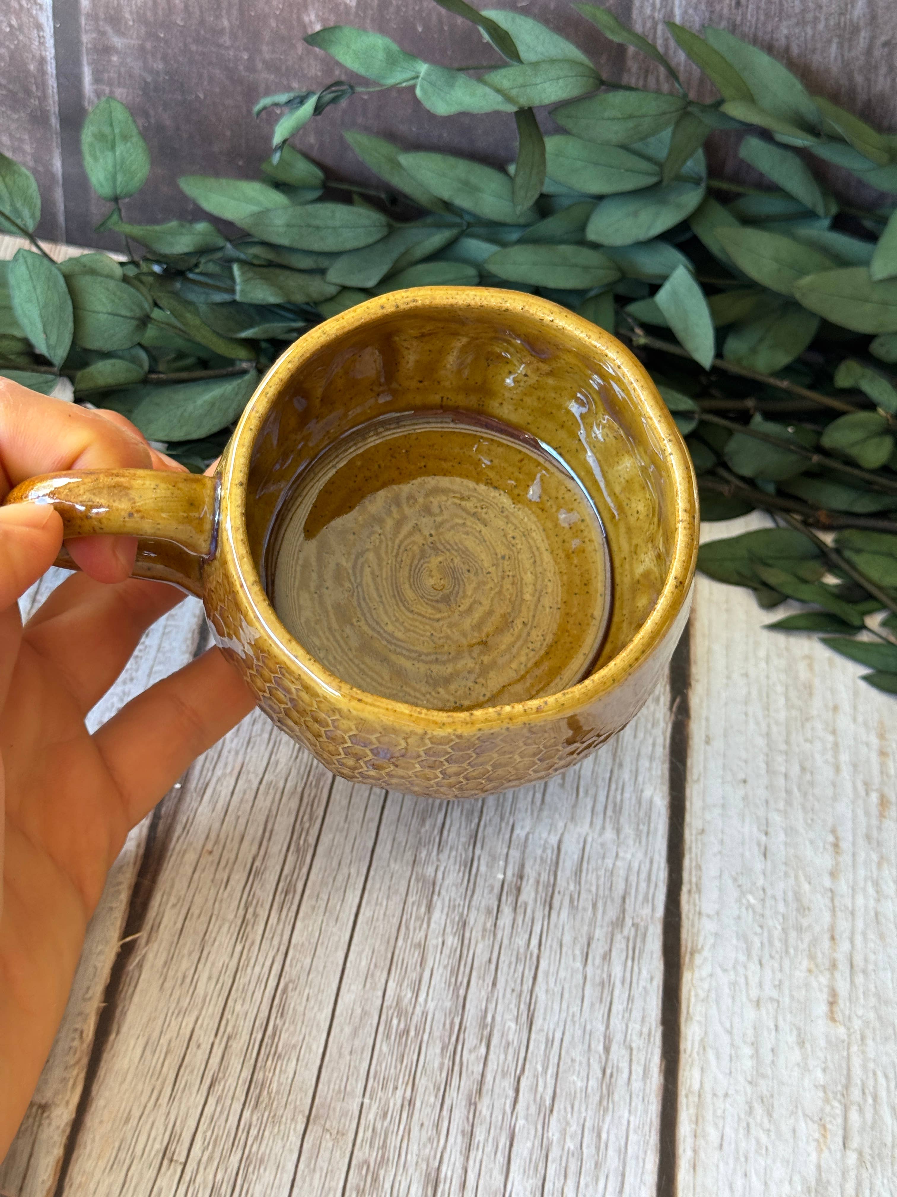 A hand holding a handmade ceramic mug with a textured mustard yellow and brown glaze, set against a rustic wooden background with green leaves.
