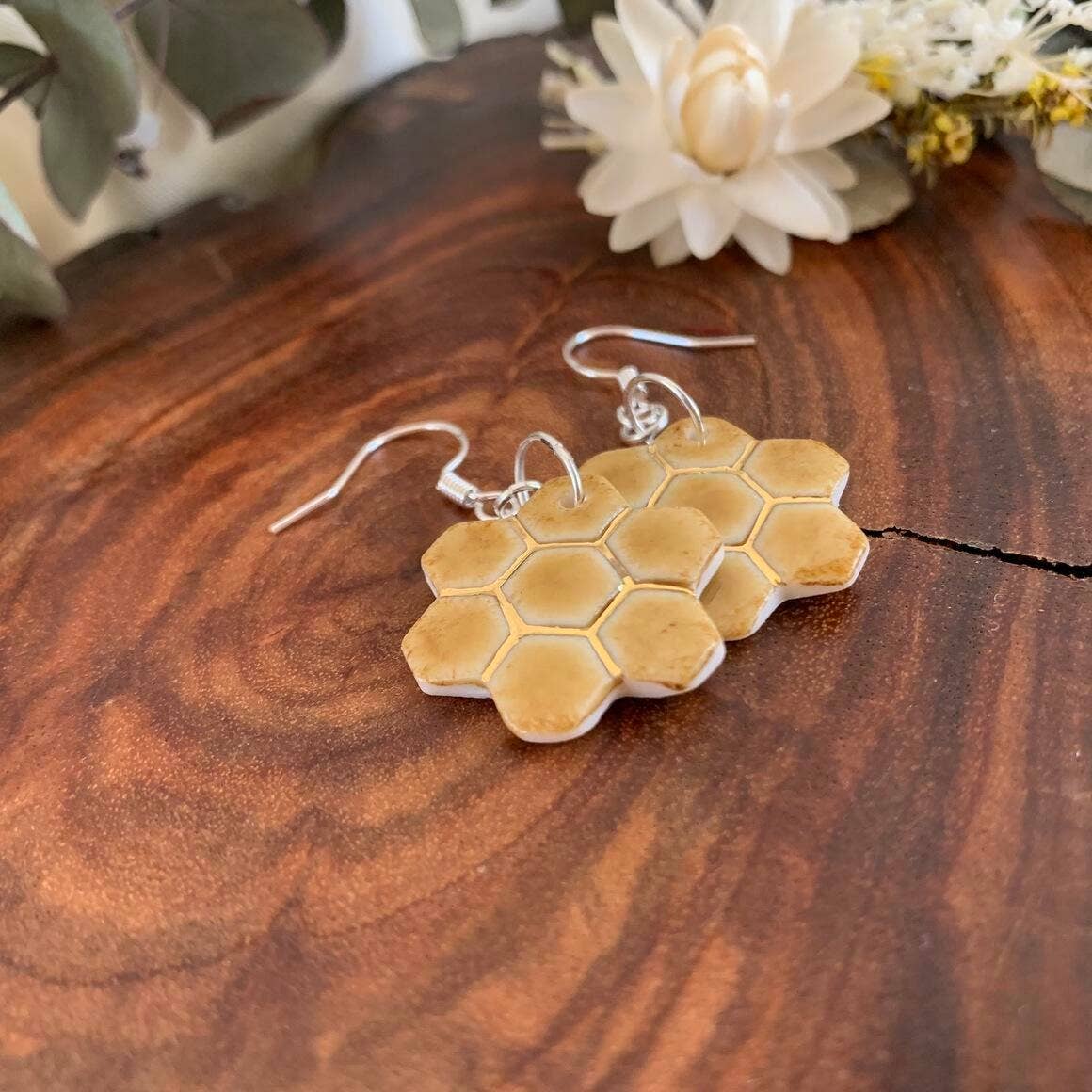 A pair of honeycomb-shaped dangle earrings with a natural tan resin body and silver fishhook wires, displayed on a rustic wooden surface with white flowers in the background.
