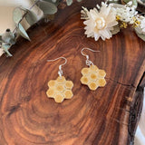 A pair of honeycomb-shaped drop earrings with a natural tan wood finish and silver fishhook wires, displayed on a rustic wooden slice with dried flowers and greenery.