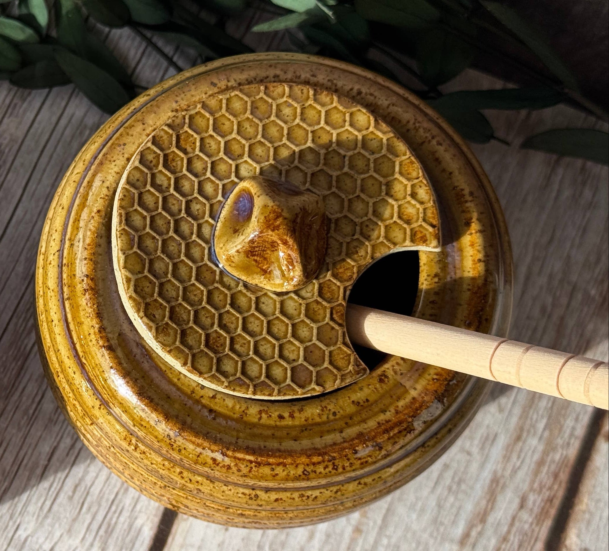 Two round ceramic honey pots with lids, featuring a honeycomb pattern and a small bee-shaped knob, displayed on a rustic wooden surface with green leaves.