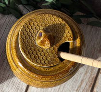 Two round ceramic honey pots with lids, featuring a honeycomb pattern and a small bee-shaped knob, displayed on a rustic wooden surface with green leaves.