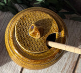 Two round ceramic honey pots with lids, featuring a honeycomb pattern and a small bee-shaped knob, displayed on a rustic wooden surface with green leaves.