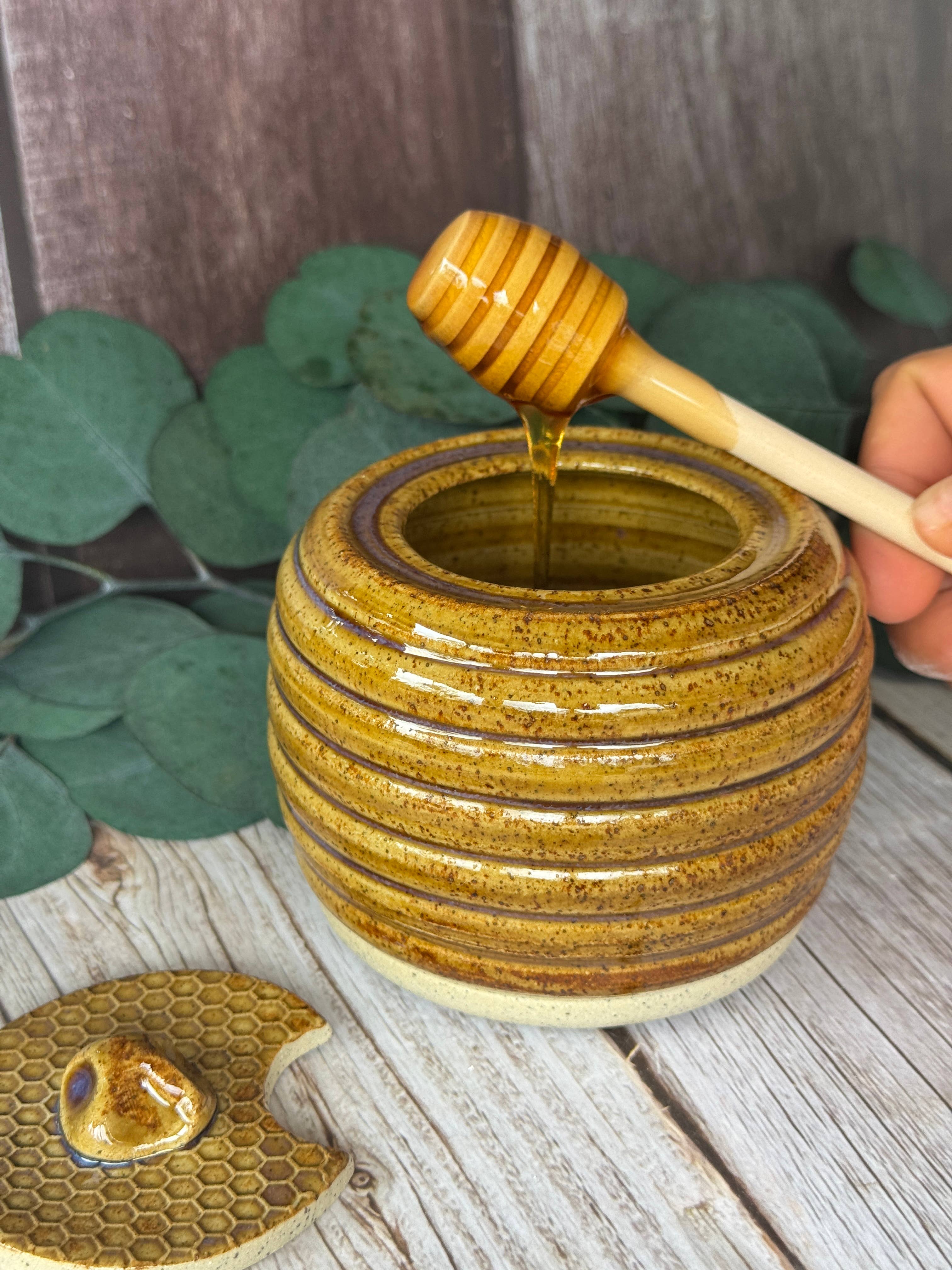A handmade ceramic honey pot with a ribbed, glazed exterior in golden-brown and amber tones, shown with a wooden honey dipper dripping honey into it. A hand holds the dipper, and a matching honeycomb-patterned lid rests on a rustic wooden surface with green leaves in the background.