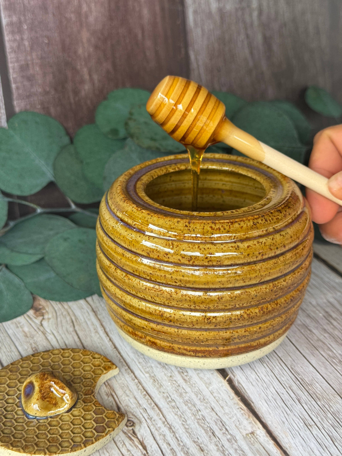 A handmade ceramic honey pot with a ribbed, glazed exterior in golden-brown and amber tones, shown with a wooden honey dipper dripping honey into it. A hand holds the dipper, and a matching honeycomb-patterned lid rests on a rustic wooden surface with green leaves in the background.