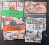 Three cookbooks laid flat on a dark wooden surface: 'Off-Grid Kitchen' , 'Natural Small Batch Cheesemaking' , and 'Sourdough Without Fail' by Kate Downham The books feature food photography and colorful title bands.