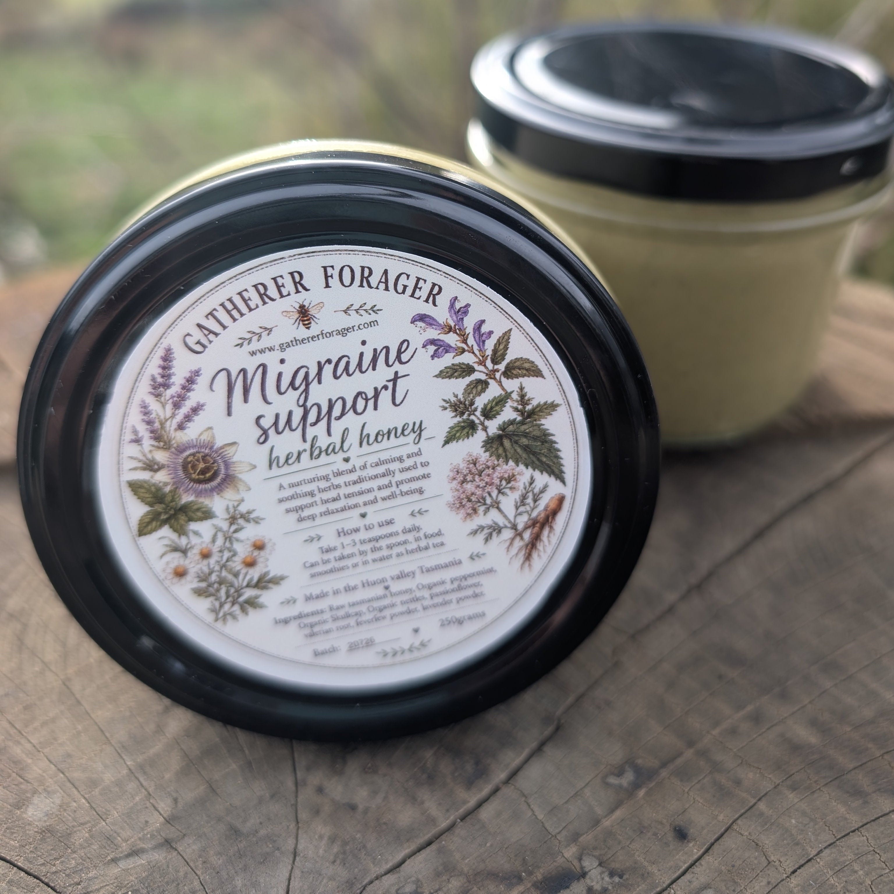 Two jars of Gatherer Forager Migraine Support herbal honey displayed on a rustic wooden surface with a blurred outdoor background.