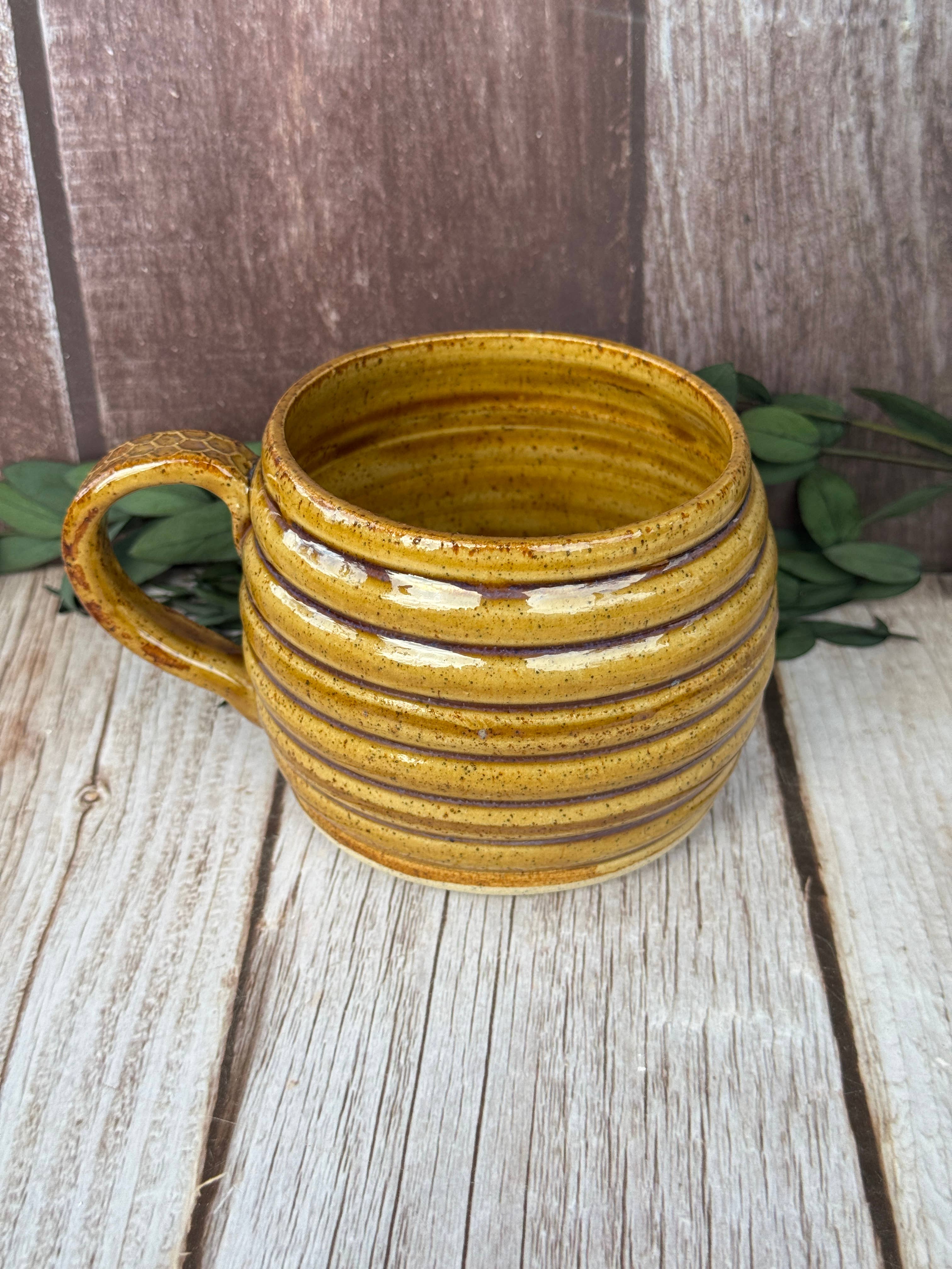 A handmade ceramic mug with a ribbed texture and a rustic mustard-yellow glaze, set on a weathered wooden surface with green leaves in the background.