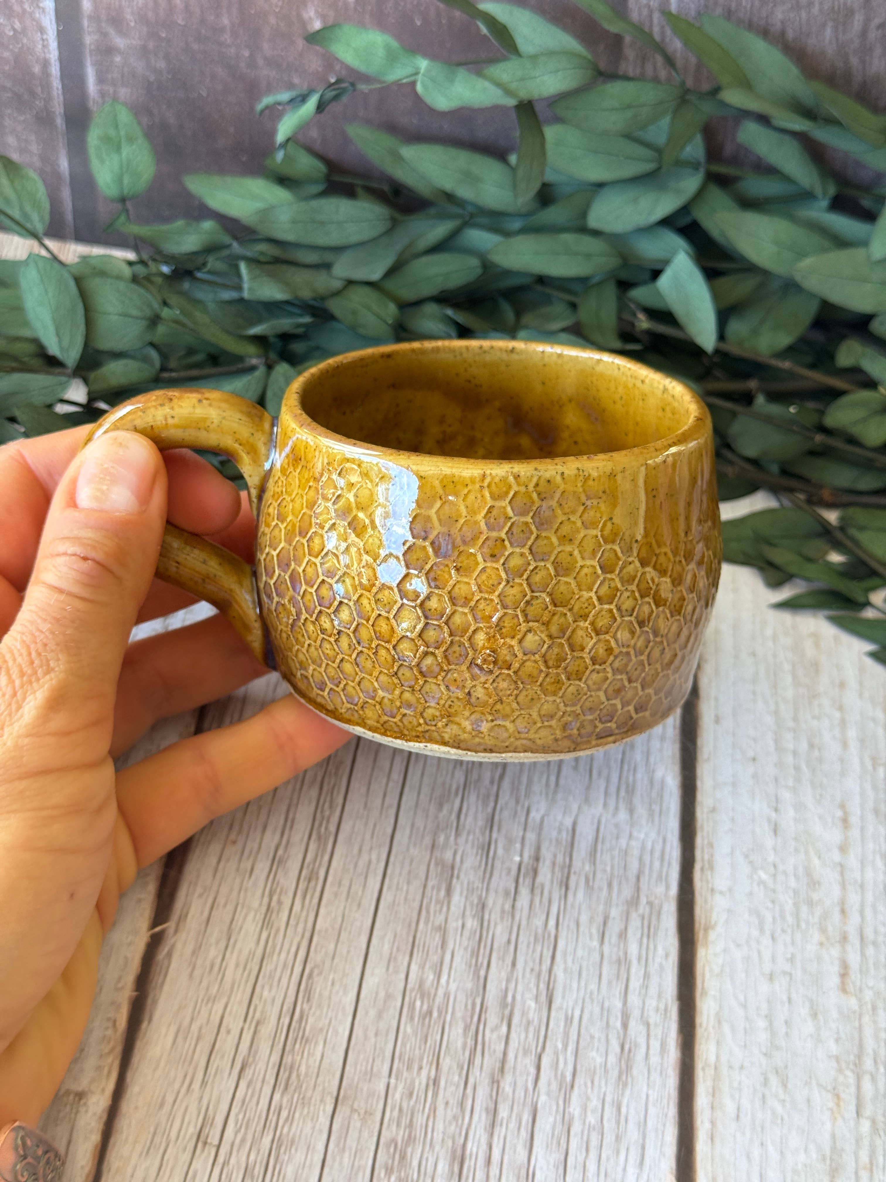 A hand holding a small ceramic mug with a textured honeycomb pattern and a glossy mustard-yellow glaze, set against a rustic wooden background with green leaves.